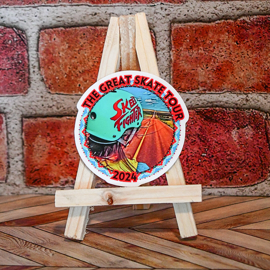 Colorful sticker with 'The Great Skate Tour' and 'Skeeter' branding on a wooden stand against a brick wall.