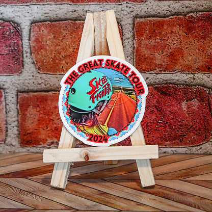 Colorful sticker with 'The Great Skate Tour' and 'Skeeter' branding on a wooden stand against a brick wall.